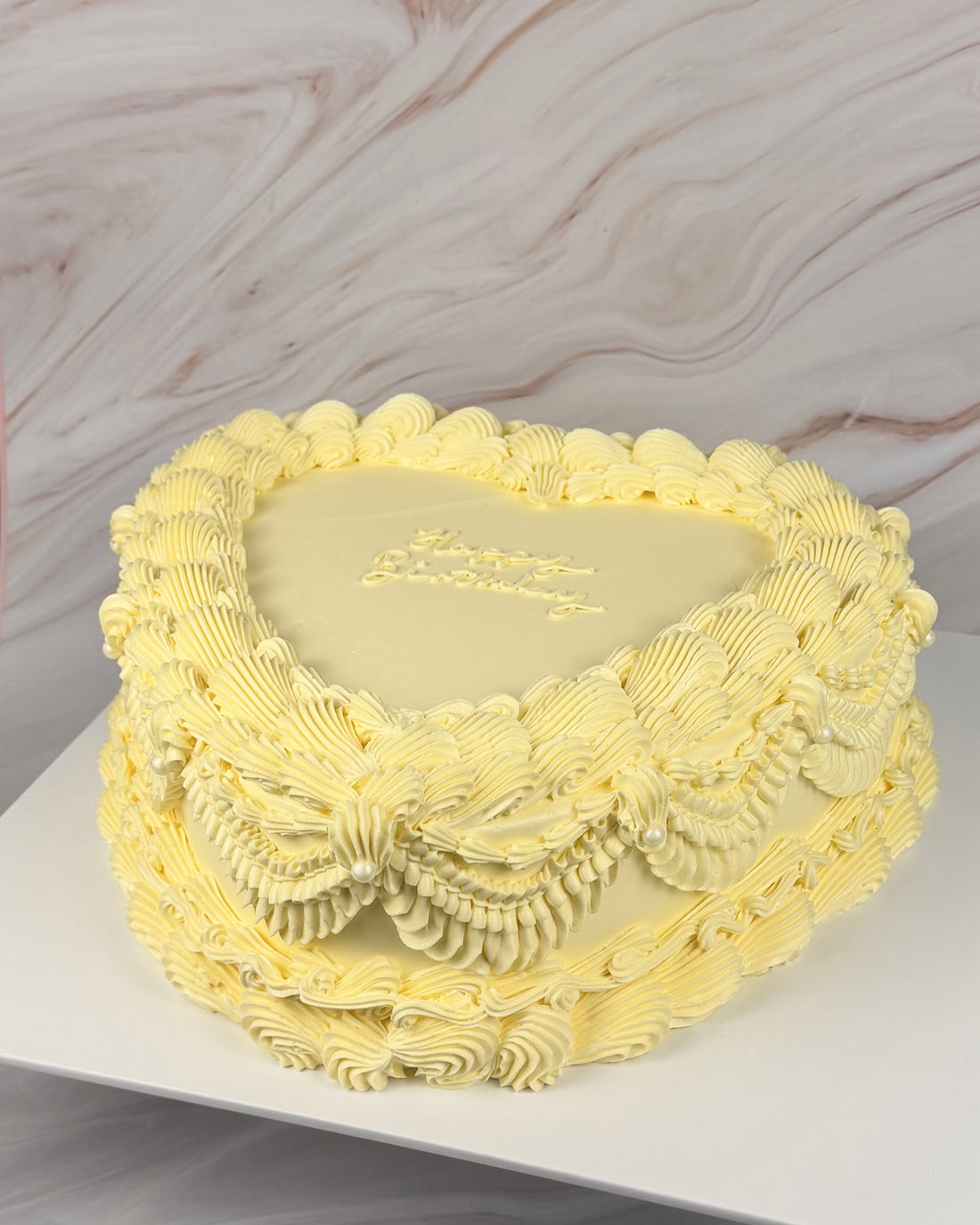 A heart-shaped birthday cake with pale yellow frosting, decorated with piped swirls and small pearl-like accents, sitting on a white board against a marble-patterned background.