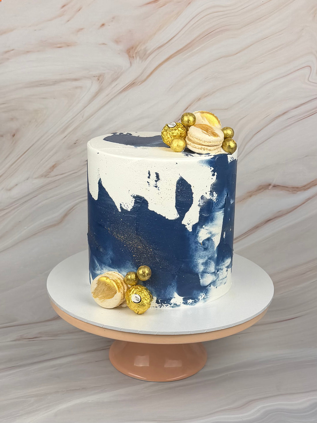 A cylindrical cake with a white and navy blue watercolor-style painted design, decorated with gold macarons and gold beads, displayed on a peach-colored cake stand against a light marble-patterned background.