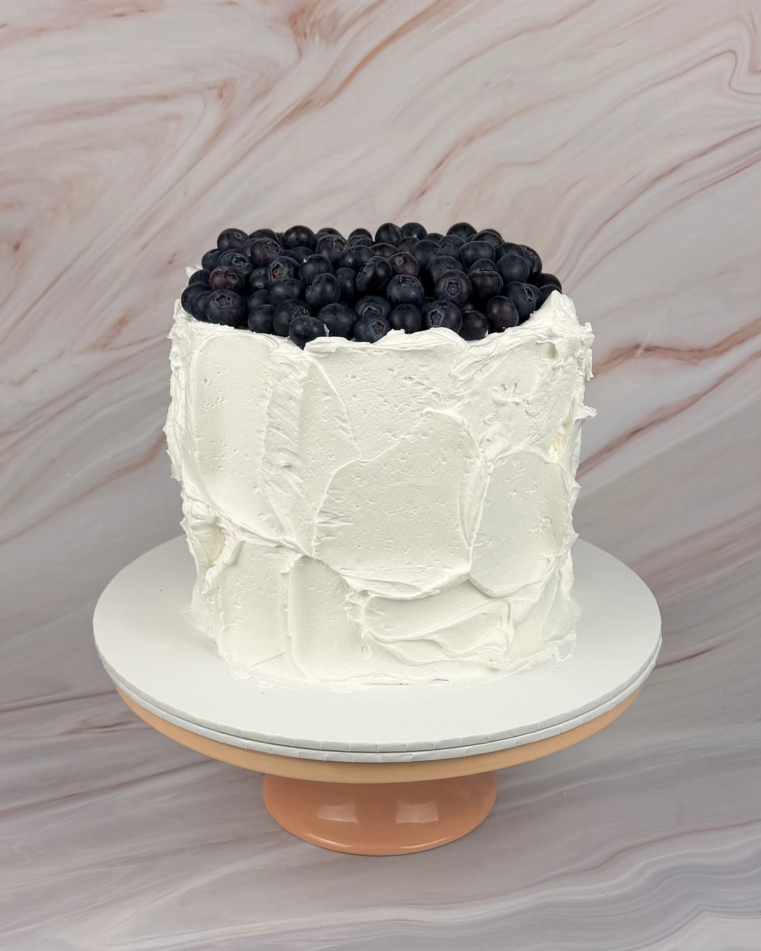 A white cake with a messy textured buttercream finish. The cake is topped with a heap of blueberries