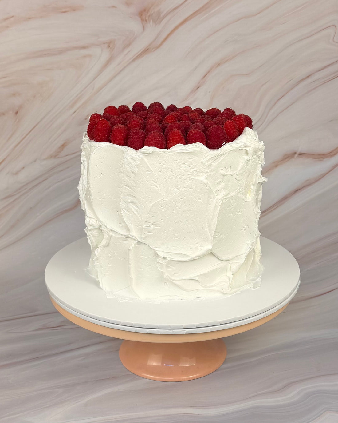 A white round cake with a messy textured buttercream finish. The cake is covered on top with raspberries.