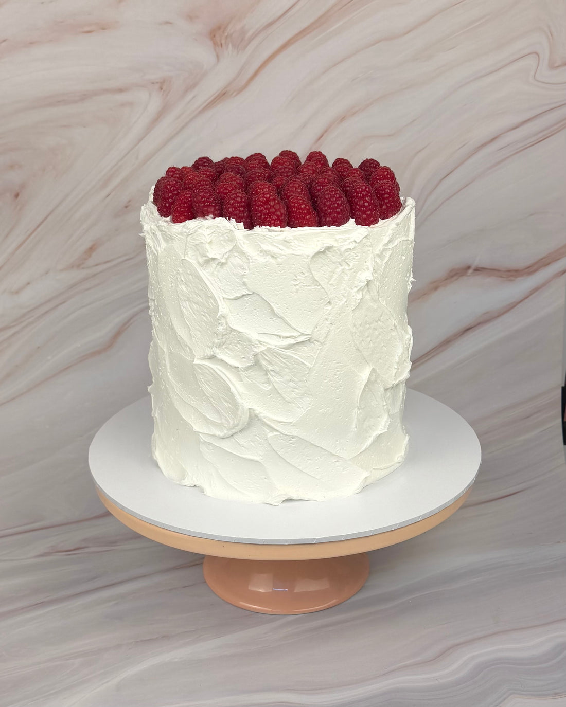 A white frosted cake topped with fresh red raspberries on top, displayed on a white round cake stand with a peach-colored base against a light marble-patterned background.